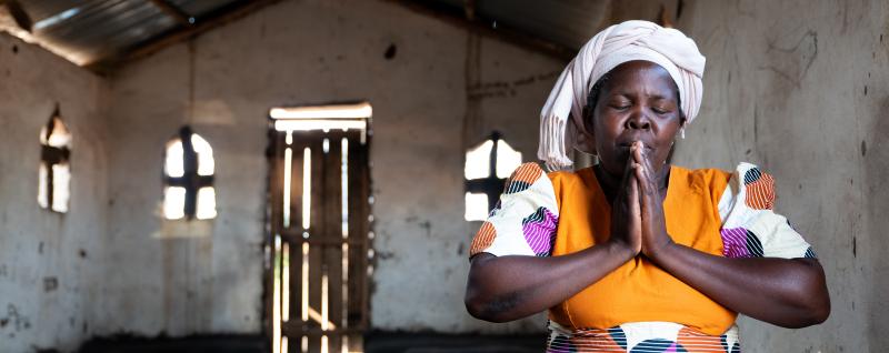 Esther sits in a church in a prayer pose with her eyes closed 